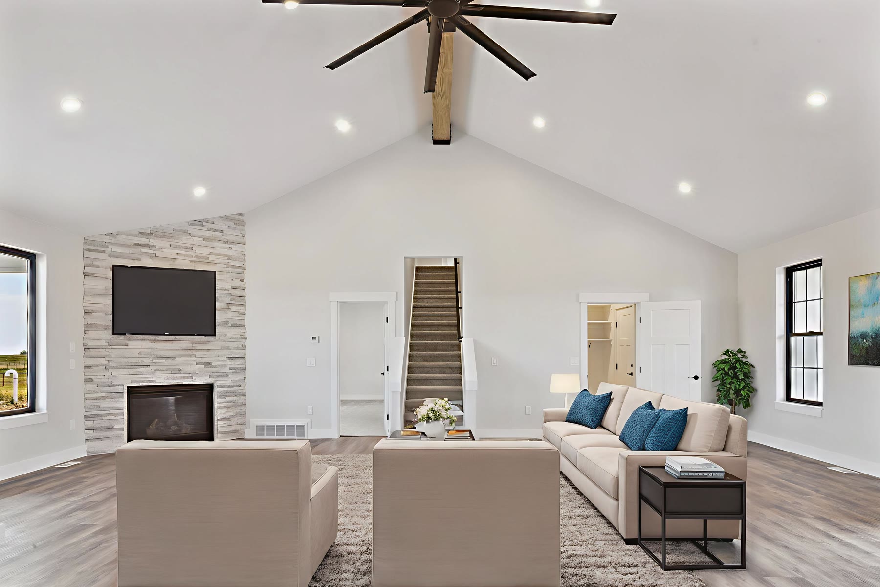 Vaulted great room in a JR Prime custom home with a wood ridge beam, stacked-stone fireplace, and black-framed windows